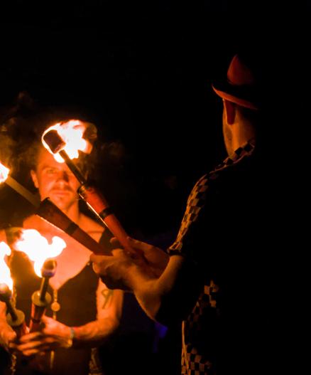 Two people, apparently male, stand holding flaming torches with a black sky behind them. One person has their back to the camera; they are wearing a trilby hat and checked shirt. The other is behind four burning torches and is wearing a unitard. They are staring at the camera. Smoke from the torches mingles with their hair, giving them an otherworldly appearance.