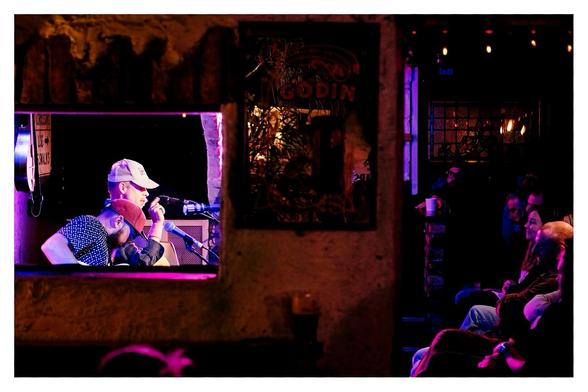 A wall separates two rooms. 

On the left is a hatch through which we see the stage area with Thomas Deakin, red trucker cap, and just beyond him, Bonnie Prince Billy performing. 

To the right is an open doorway to the same room. There we see their audience.