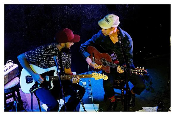 Seen from above, Thomas, red hat on the left playing guitar, leans in towards Will Oldham, white cap, facing Thomas, also playing guitar.
