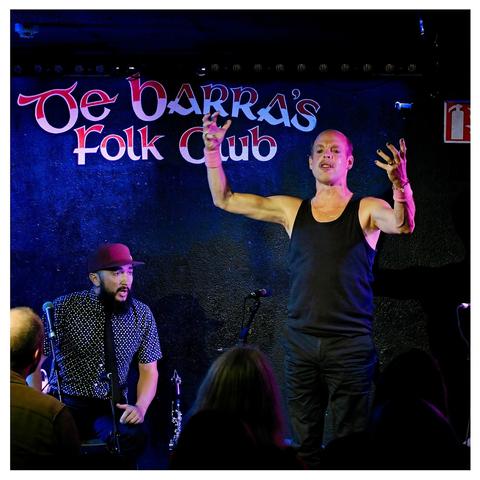De Barra’s Folk Club is written on the wall behind them.

Thomas seated on the left.
Will Oldham, Bonnie Prince Billy, stands, now, in a tank top, without his cap, his arms and fingers lifted, lifting us in song.
