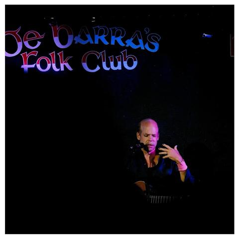 A square photo. 

Top left De Barra’s Folk Club written on the wall. 

Lower right Will Oldham, Bonnie Prince Billy, no cap, seated at the mic, his left hand raised, fingers spread in gesture.

The rest of the photo is in darkness. 

One of those nights where friends, old and new have gathered together under stormy skies.