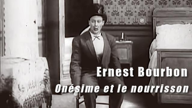 Capture d'écran horizontale du film « Onésime et le nourrisson » de Jean Durand avec Ernest Boubon, sorti le 23 août 1912. Onésime en costume, chemise blanche, avec chapeau haut de forme sur la tête est assis sur une chaise au centre de l'image dans une chambre à couché, lit sur la droite de l'image, chevet au fond et au centre derrière Onésime, à côté du chevet une armoire avec miroir, une table avec une nappe tissée sur la gauche. Entre la table et l'armoire de longs rideaux blanc d'une fenêtre.