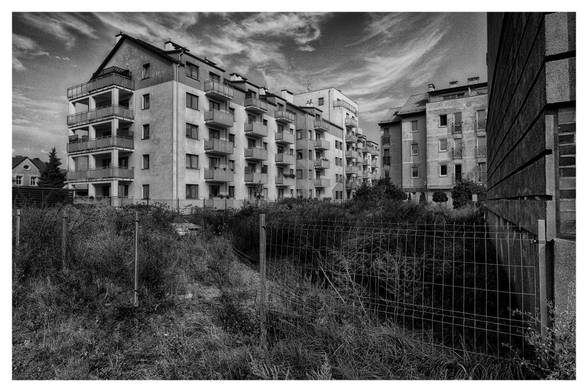 Wohnanlage mit umzäunten verwildertem Grundstück im Vordergrund, auf dem vor längerer Zeit ein Pkw abgestellt wurde