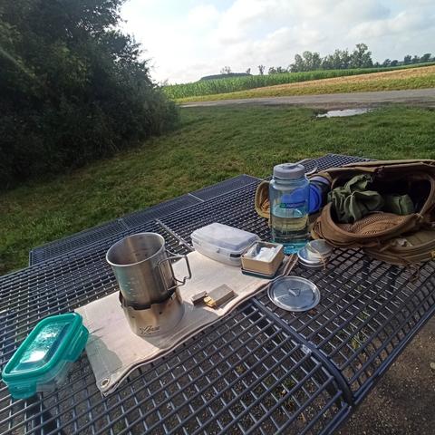 Ein Tisch auf dem ein geöffneter Rucksack liegt. Im Vordergrund ein X Boil Spirituskocher, mit einer Edelstahltasse. Im Hintergrund ist ein Gebüsch und ein Blick auf ein Feld.