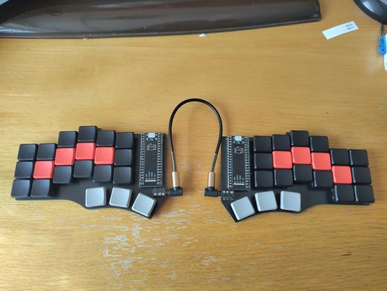 An assembled Cantor keyboard. It is a split keyboard with an ergonomic 3 by 6 design and with 3 thumb keys. The homerow has red keycaps, the thumb keys are grey. All other keycaps are black