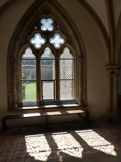 Ein mittelalterliches Fenster mit Spitzbogen in einem Kreuzgang, von drinnen nach draußen fotografiert. Das Fenster ist im größeren unteren Teil dreiteilig mit hohen Bleiglasfenstern mit kleineren Spitzbogen, oben sind im Dreieck drei kleinere Fenster in Form von Kleeblättern. Draußen erkennt man weitere Mauern und Rasen. Die Sonne scheint durch die Scheiben und wirft die Form der Fenster auf den Steinboden im Inneren.