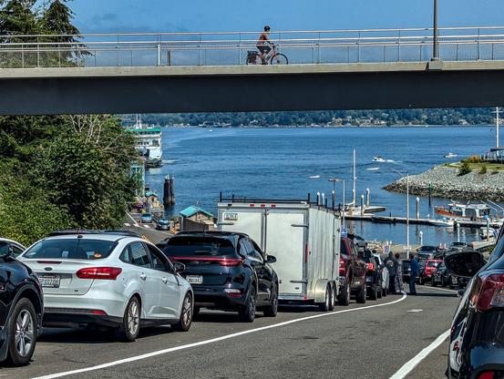 Photo shows the Point Defiance ferry dock and marina. Cars are lined up awaiting the ferry. A cyclist rides across a pedestrian bridge.