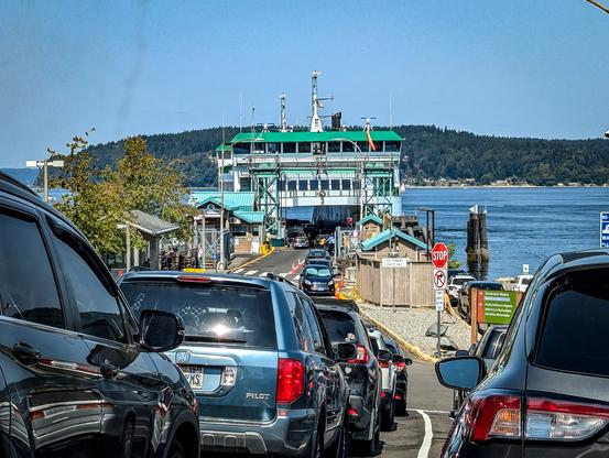 Photo shows the Point Defiance ferry dock with a ferry ready to unload cars. Cars are lined up awaiting the ferry.