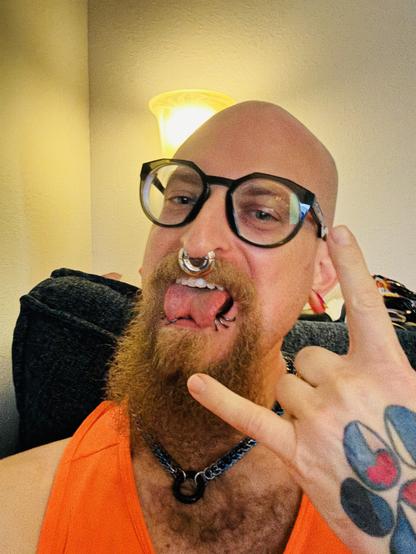 A person with a shaved head and a full goatee holding their hand in the “horns” symbol, sticking out their split tongue, with a heavy gauge septum ring and multiple lip rings. They are wearing large glasses and an orange tank top, with visible tattoos on their hand.