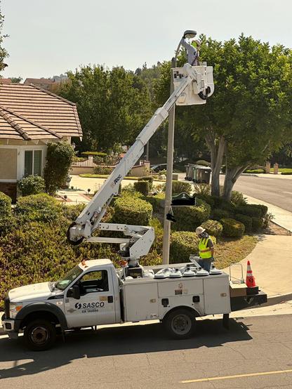 A utility truck with a raised bucket is seen near a residential area. A worker in a yellow safety vest and helmet is in the bucket, performing maintenance on a utility pole. He is replacing the streetlight element. The truck below has many more replacement lights visible.