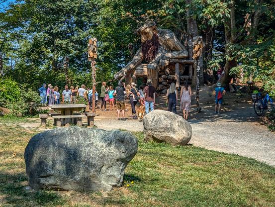 The photo shows a large wooden sculpture of a troll in a wooded setting. People are gathered around the sculpture to view it. Two large stones are in the foreground, likely glacial erratics left from the last ice age.
