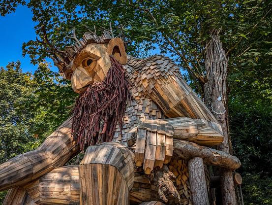 The photo shows a large wooden sculpture of a troll in a wooded setting. The sculpture is made from recycled wood.