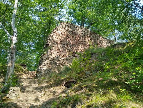 Ruiny kamiennego muru na zboczu w słońcu. Mur wykonano z różnobarwnych kamieni — dostrzec można odcienie szarości, piasku i pomarańczu. Wzdłuż muru zbocze po prawej stronie porośnięte jest trawą i inną roślinnością. Po lewej stronie na wprost zmierzają szare, kamienne schody w górę. Obok nich, białobrązowe drzewo. W tle zielone korony drzew, przez które prześwituje błękitnobiałe niebo.