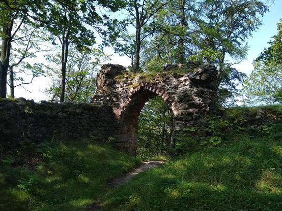 Kawałek muru z portalem w zacienionym zakątku wzgórza. Po bokach pozostał niższy fragment muru, przy portalu trochę wyższy. Portal ma kształt zakończonego spiczasto łuku, wyłożonego od środka poprzecznymi cegłami. Po prawej stronie mur zasypany ziemią i porośnięty roślinnością. Do portalu prowadzi wydeptana ścieżka, dookoła trawa. Po drugiej stronie widać drzewa, kilka wyrasta powyżej muru, jednak tu są niezbyt gęste, jakby ciut rozmyte na tle prześwietlonego białobłękitnego nieba.