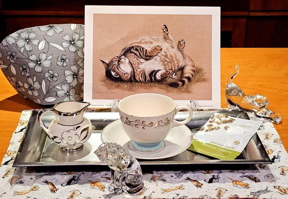A tea tableau in muted, almost sepia, tones with a pen & ink drawing of a fat tabby cat on its back with feet in the air. There are also two Lenox crystal cat figurines, one sitting and grooming its paw, the other stretched sinuously in a bowing position. There is vintage MidCentury china on a silver tray, a grey & white floral cozy, a cat-patterned placemat and the tea is Jasmine Pearls green tea from Adagio