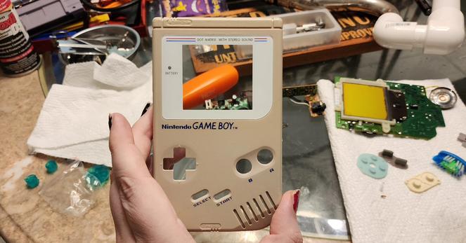 In focus is Erika's hand holding a front half of a Nintendo game boy shell with a brand new white screen protector. Off to the side on a messy table are the hardware internals from the system