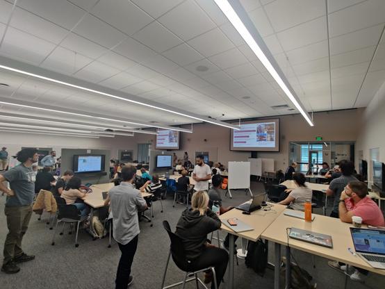 Closing session of the 2024 NASA-NSF Earth Sciences UW Hackweek! Lots of people around tables looking to the podium where the organizers of the ICESat-2, SnowEx and GeoSMART tracks stood and were giving closing remarks.