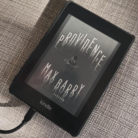 An e-reader displaying the book "Providence" by Max Barry. The device is connected to a charger and placed on a textured surface.