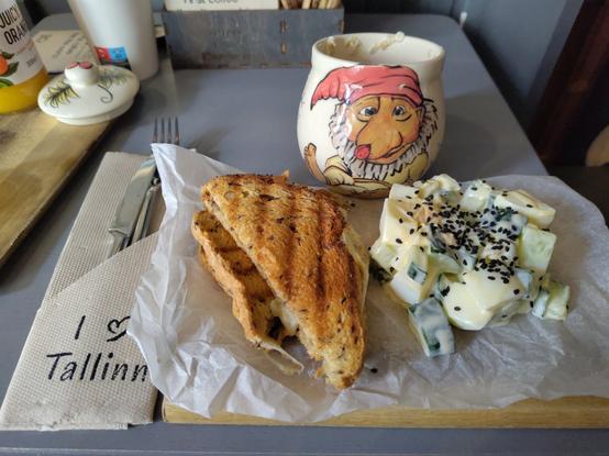 A toast and cucumbers in mayonnaise, and next to the plate a napkin with the text: “I ❤️ Tallinn”. In front of the plate a cup with a gnome painted on it.