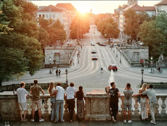 On a summer evening in Munich, locals and tourists alike pause to admire the sunset from the vantage point near the Friedensengel. The golden light spills across the city, highlighting the architecture and the lush greenery of the surrounding area, while people share a moment of tranquility above the bustling street below.