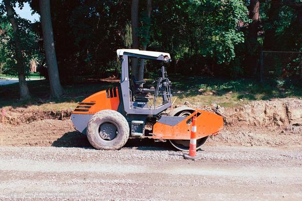 A photograph of an orange and grey roller compactor vehicle with a white roof. It is sitting on gravel in front of disturbed brown dirt and some trees. An orange-and-white-striped traffic cone is in front of it. Labels on the compactor read:IH7 R4-15 and COMPACT Line.