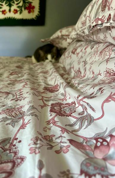 Looking across a made bed. The foreground is mostly a white duvet with pink and gray flowers. At the far side of the bed is a blurry, piebald tabby cat with big eyes crouches beneath the edge of a pillow. In the background is a blue wall with a colorful hanging from Hawaii of quilted hibiscus.