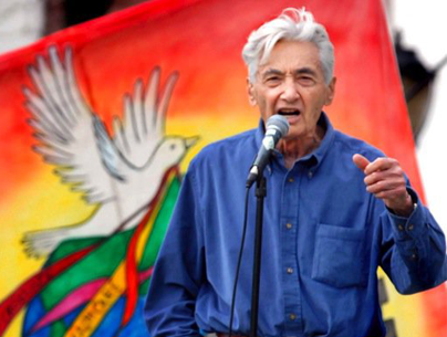 Howard Zinn, with gray hair and a blue button down shirt, speaking at a microphone, with a colorful flag in the background that has a peace dove on it.