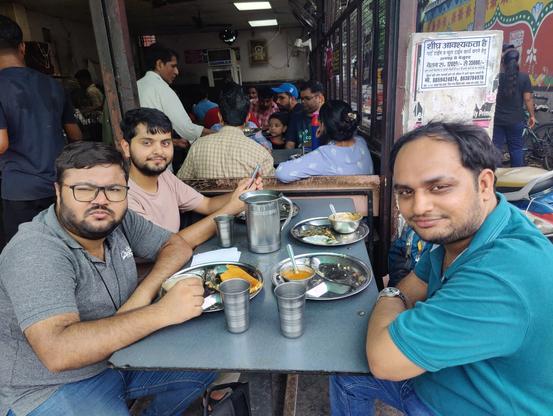 Three folks at lunch table looking at the camera. More people can be seen in the background.
