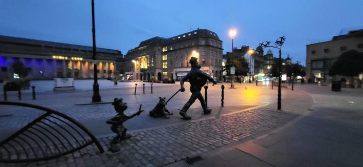 Colour photo of Desperate Dan, Dawg and Minnie The Minx statues in Dundee, Scotland. These are characters in the comic The Dandy  which originated Dundee, first published in 1937. Photo shows characters from behind with historical architecture buildings in background. A clear blue evening sky says Beanotown wants you back soon..