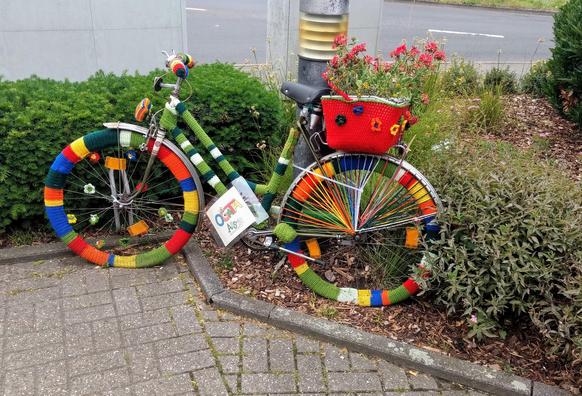 Fahrrad, das komplett eingehäkelt ist. Alle Farben des Regenbogens sind vertreten, bunt gestreift. Sogar ein gehäkelter Korb steht auf dem Gepäckträger. 

Das Rad lehnt an einen Laternenpfahl und steht zur Hälfte in einer städtischen Grünfläche.