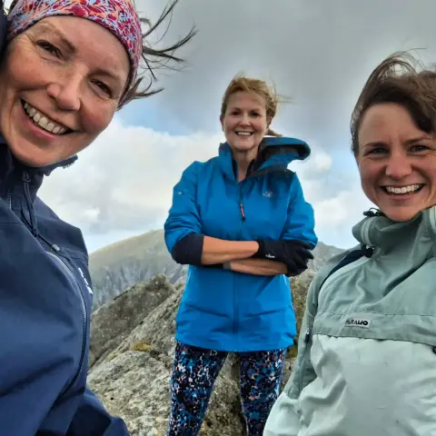 A summit selfie of threevwomen on the top of Cir Mhor. They are grinning and the wind is blowing their hair around.