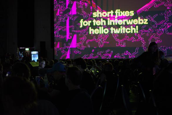 big main display says "short fixes for teh interwebz, hello twitch!". underneath we see rows and rows of people at computers in the dark