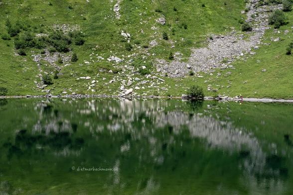 Das Foto zeigt einen ruhigen Bergsee mit spiegelglattem, grünlichem Wasser, in dem die umliegende Landschaft reflektiert wird. Am gegenüberliegenden Ufer erstreckt sich eine grasbewachsene, steinige Bergflanke. In der Ferne, am Ufer des Sees, sitzen zwei Personen in bunter Kleidung, die klein im Verhältnis zur Landschaft wirken. Der Hang ist durchzogen von kleineren Felsen und Büschen, und die Szenerie wirkt insgesamt ruhig und abgeschieden.