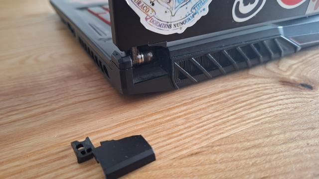 Zdjęcie tyłu laptopa, dziura w obudowie i ułamany element na stole.