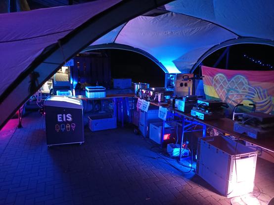 Pavillon mit Waffel Regenbogen Flaggen, Gefriertruhe mit der Aufschrift "Eis", und verschiedenstem Waffel zubereitungs Geraffel auf Bierbänken.

bei nacht, mit buntem licht beleuchtet.

Dank an glowl