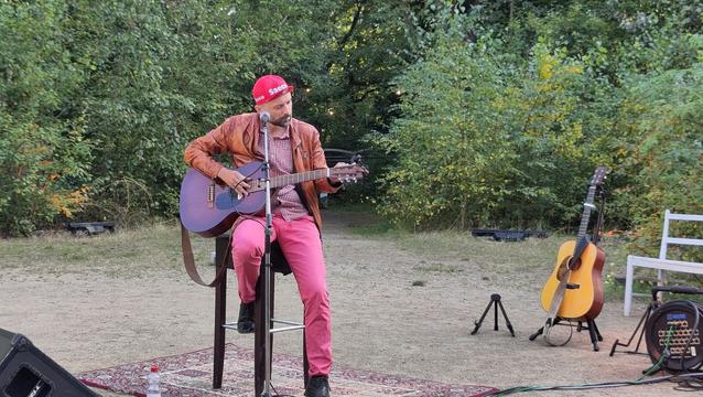 BooHoo, mit roter Mütze, brauner Lederjacke und Gitarre auf einem Hocker. Open Air. Vor Gebüschen.