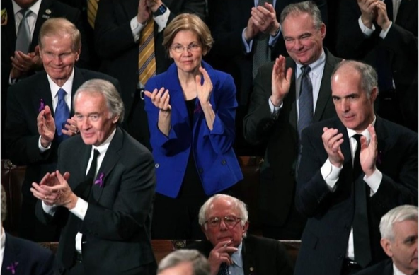 A picture from one of trump's speeches during his presidency showing a shot of the Democrat's side. 

All Democrats shown are standing and applauding except for one person sitting down, hand on his chin, with a not-pleased look on his face. 

Bernie Sanders