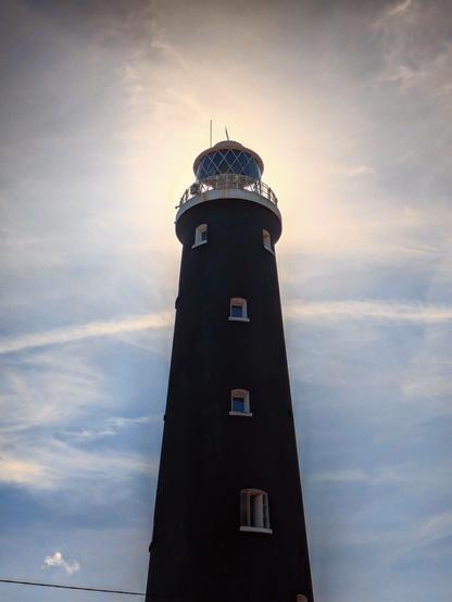 An image of a black coloured lighthouse. The sun is shining from just behind the top of the lighthouse. The rest of the sky is blue and wispy white clouds.