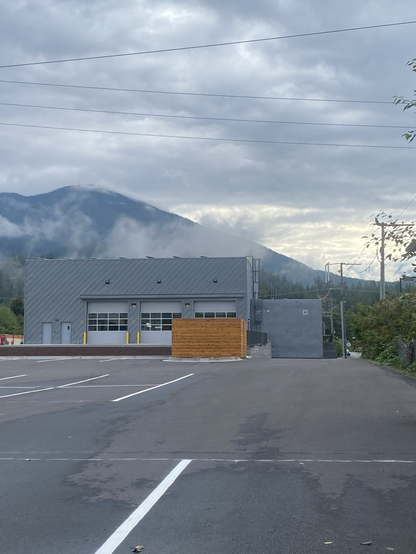 The view of a new fire hall from the back side of the building with clouds and a mountain in the background while the foreground features a paved parking area.