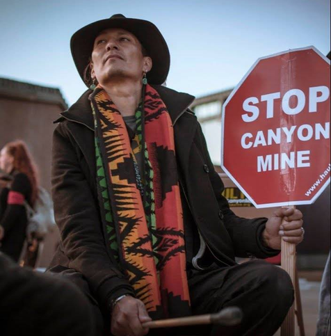 A photograph of a man, wearing a brown hat and jacket, with an orange, red, amber and green striped scarf with black designs. The man is wearing earrings, and looking up. He holds a stop sign that reads: STOP CANYON MAINE. With a link to HaulNo.com