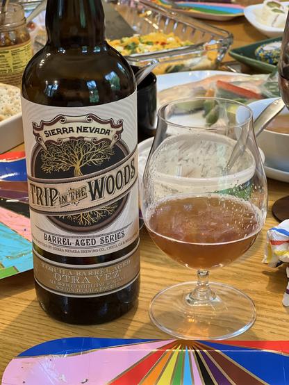 A 750ml bottle of Sierra Nevada Trip in the Woods barrel-aged series stands in front of a table covered in signs of a party. A snifter glass with a bit of the dark golden beer is to the right