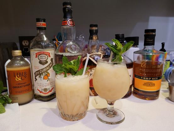 Two Mai Tai cocktails, garnished with mint, set in front of bottles of ingredients: orgeat, orange curaçao, and four rums.