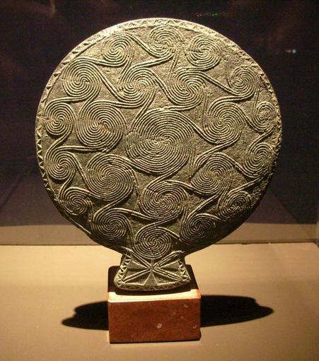 Oggetto piatto e tondo nel contesto di un museo, collocato in posizione eretta sopra un piedistallo. Spirali decorano la sua superficie lievemente convessa.