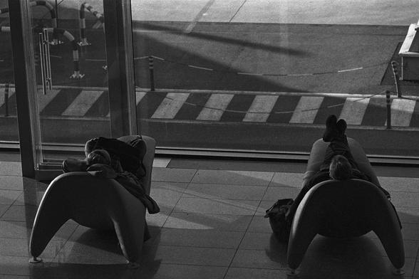 The image shows two people lying on lounge chairs, facing towards large windows. The view through the windows includes a road with painted stripes and some outdoor structures. The lighting suggests it might be early morning or late afternoon, casting shadows on the ground inside. Each person has a bag placed near them, indicating they might be travelers waiting in an airport or other transportation hub.