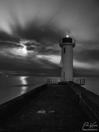 Photo, en format vertical, en noir et blanc, de nuit, présentant un phare au bout d'une jetée sous un ciel partiellement nuageux et la lune éclairant la mer entre les nuages. Un nez a été rajouté à la tour du phare, faisant penser un peu à un moaï de l'île de Pâques.