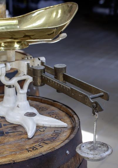 An antique balance scale with a shiny brass bowl, white-painted base, and two metal weights. The scale is placed on a wooden barrel with visible markings.