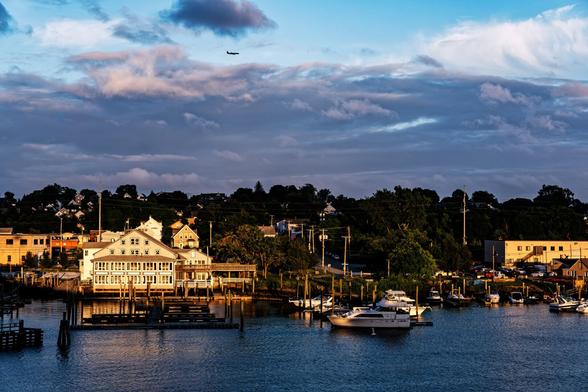 A color photograph of the opposite bank of a river. Late day sun is shining on the buildings, docks, and boats along the riverbank, while the leaves on the trees higher up on the hill are shadowed. Grey and white clouds fill the much part of the sky, and where it is blue closer to the top of the frame, an airliner is passing, left to right.