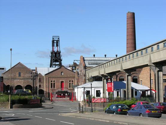 The National Mining Museum Scotland at Newtongrange. The image shows a view across a road with a car park on the right. A range of large brick colliery buildings is topped off by the winding gear supported on a black metal structure just to the left of the centre of the frame, while a tall brick chimney is on the right of the frame, above the housing for a conveyor system. The sky is almost completely blue.