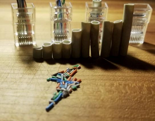 An art piece composed of cutoffs from RJ45 connectors, arranged on a wooden surface.

In the background, four connectors stand on end, evenly spaced. Three have scraps of cables trapped inside and dangling above them. The last is empty, abandoned without any wires inside.

Before them, ten pieces of cable tubing stand vertically on end, side by side, arranged from shortest to longest in a shape resembling the signal strength icon.

In the foreground, a colorful jumble of tiny cut wires form a thunderbllt shape, similar to the electric emoji.