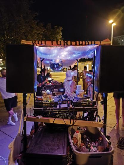 Eine Fahrrad-Plattform von hinten. Es sind zwei große Lautsprecher zu sehen und ein DJ-Pult mit Mischpult und CD-Playern. Darunter ein Behältnis mit verschiedenen Getränken. Am Dach ist eine Aufschrift "Kulturmobil" zu sehen.